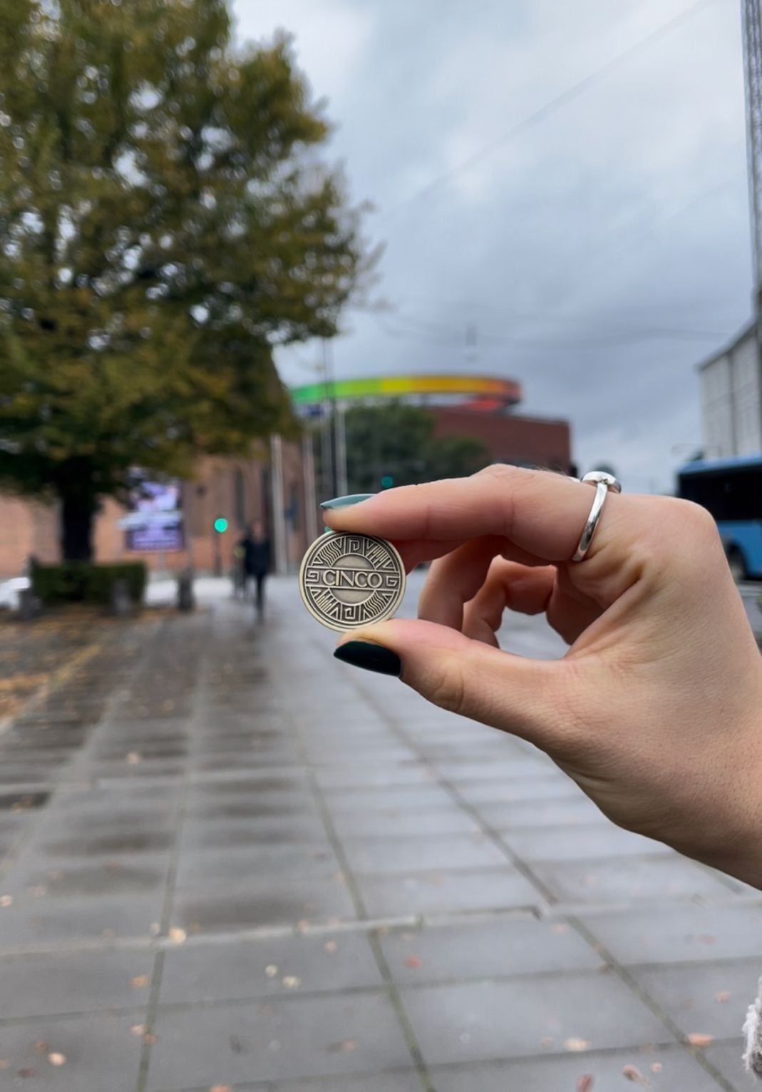 En hånd holder en guldmønt med Cinco-logoet foran ARoS og regnbuepanoramaet i Aarhus