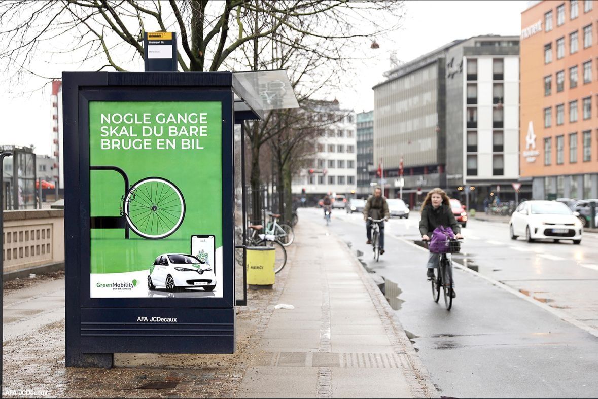 GreenMobility-plakat i et busskur i København: "Nogle gange skal du bare bruge en bil" – et cykelhjul i et cykelstativ og en hvid elbil, mens cyklister kører forbi