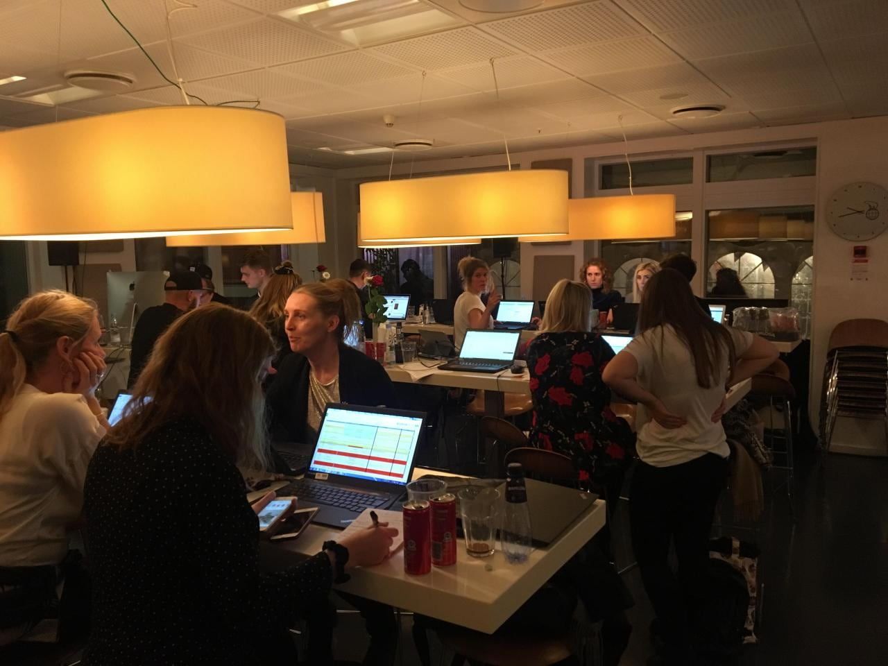 Et fyldt lokale om aftenen, hvor holdet sidder ved borde med bærbare computere under store lamper